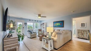 A living room with a sofa, a television, a ceiling fan, and a bookshelf. - Tide Dyed – A Colorful Coastal Escape (Topsail Beach)