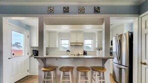 A kitchen with white cabinets, a stainless steel refrigerator, and a bar area with stools. - Tide Dyed – A Colorful Coastal Escape (Topsail Beach)