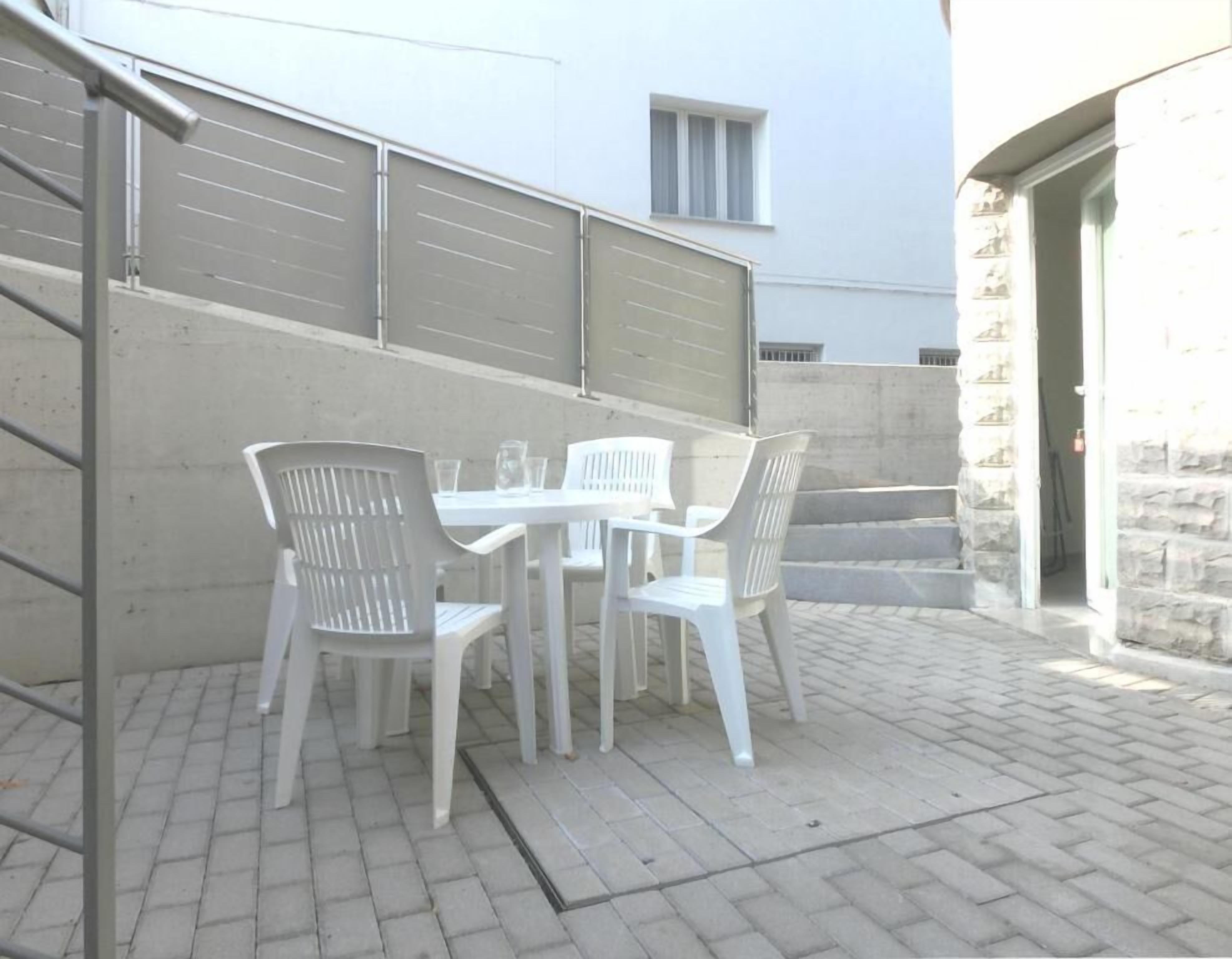 Une terrasse avec une table et des chaises, un mur en béton et un bâtiment en pierre en arrière-plan.