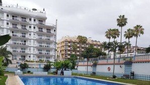 Une piscine dotée d’une terrasse carrelée, entourée de verdure et de palmiers, avec des immeubles à plusieurs étages en arrière-plan.