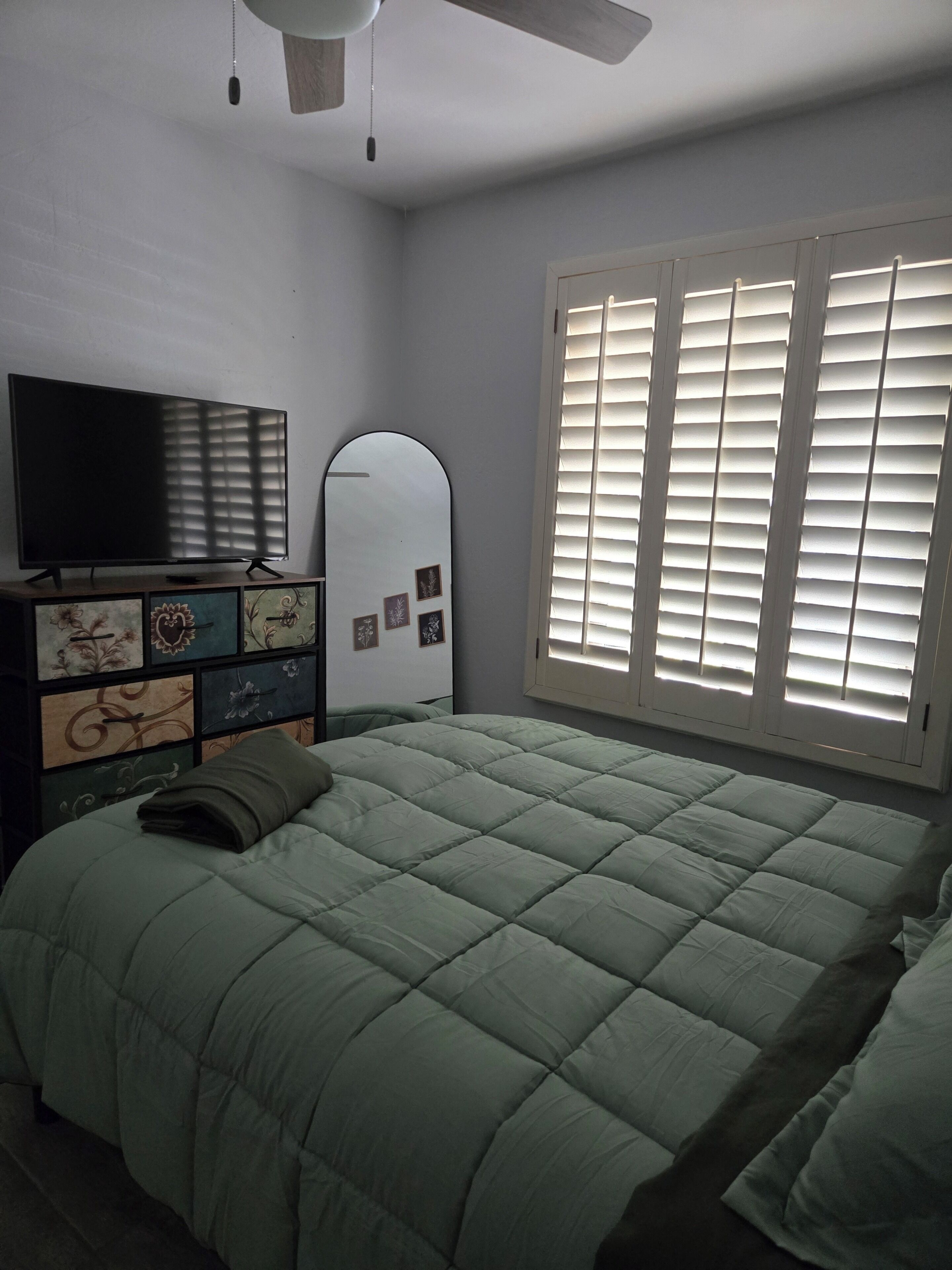 Une chambre à coucher comprenant un lit, une commode ornée de panneaux décoratifs, un téléviseur, un ventilateur de plafond et une fenêtre avec des stores.