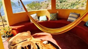 A hammock in a room with a view of the ocean and hills, a wooden sculpture, and a red cushioned bench. - Villa Xcaanda (San Pedro Pochutla)