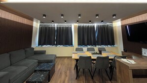 A modern room with a sofa, dining table, chairs, a TV, and large windows with curtains.