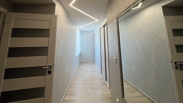 A narrow hallway with a wooden floor, a ceiling with linear lighting, and a mirrored wall on one side.