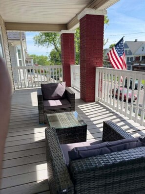 A covered porch with wicker furniture, a white picket fence, and an American flag. - NEW! Modern Niagara Loft with Private Sunset Balcony (Niagara Falls)