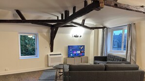 Ein modernes Wohnzimmer mit freiliegenden Holzbalken, einem grauen Sofa, einem Flachbildfernseher und einem Fenster mit Blick auf Gebäude.