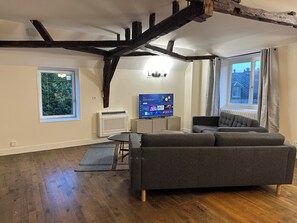 A modern living room with exposed wooden beams, a grey sofa, a flat-screen TV, and a window with a view of buildings. - Private Apartment - T3+T4 - 190m² - 10 pers - Courtyard&Parking - Hypercentre Bourges (Bourges)