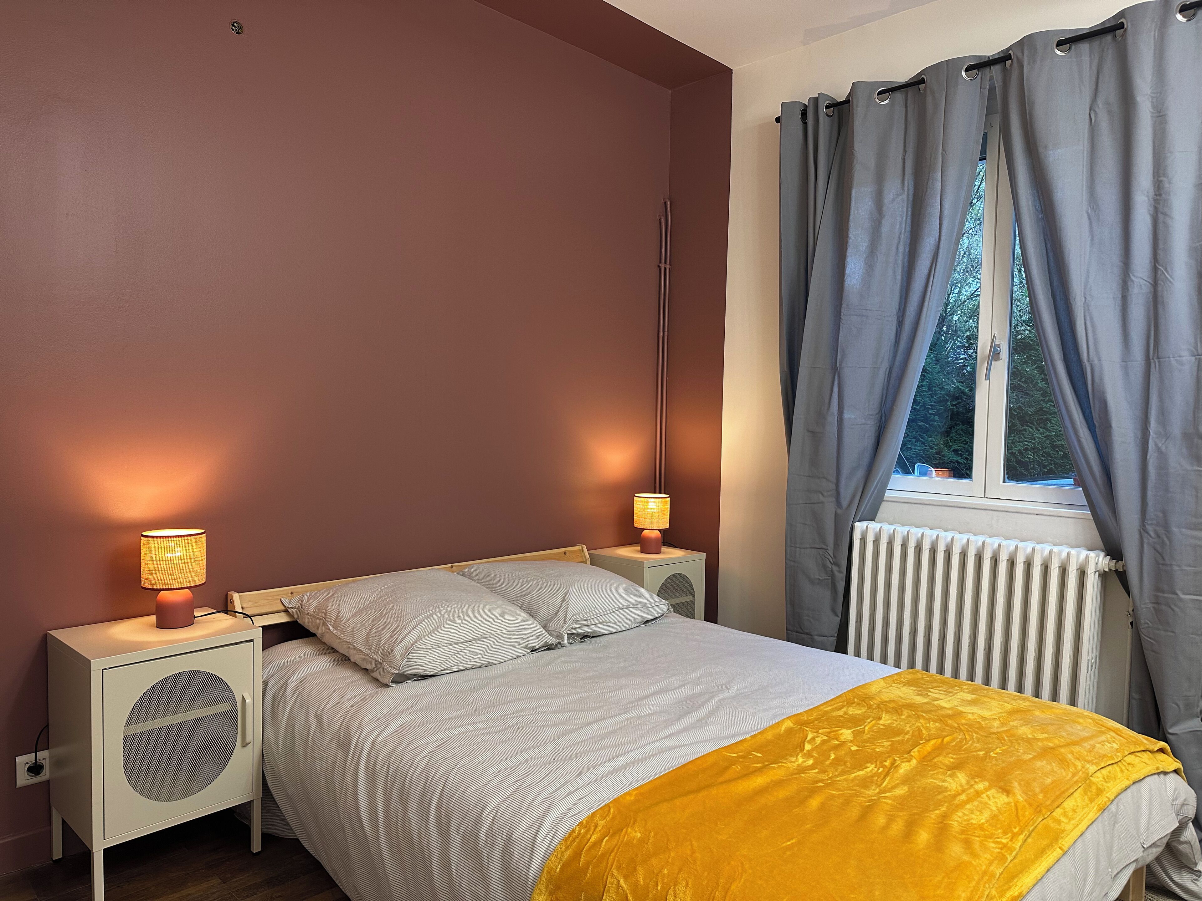 A bedroom with a bed, two bedside lamps, a radiator, and a window with curtains.