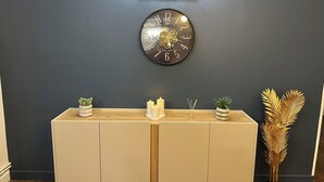 Eine wandmontierte Uhr mit dunklem Zifferblatt und goldenen Zahlen, ein heller Konsolentisch mit Zierpflanzen und eine Wandleuchte mit drei Lampen.