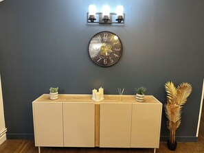 A wall-mounted clock with a dark face and gold numbers, a light-colored console table with decorative plants, and a wall sconce with three lights. - Private Apartment - T3+T4 - 190m² - 10 pers - Courtyard&Parking - Hypercentre Bourges (Bourges)