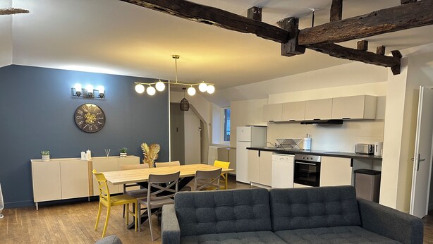 Ein modernes Wohnzimmer mit grauem Sofa, einem Essbereich mit gelbem Sessel und einer Küche mit weißen Schränken und einem schwarzen Backofen.