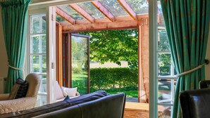 Une pièce ouverte donnant sur un jardin grâce à de grandes portes vitrées, avec une structure de plafond en bois et un canapé agrémenté de coussins.