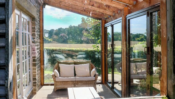 Un espace extérieur aménagé, abrité, avec un canapé en osier et des coussins, une table en bois et une porte en verre donnant sur un jardin.