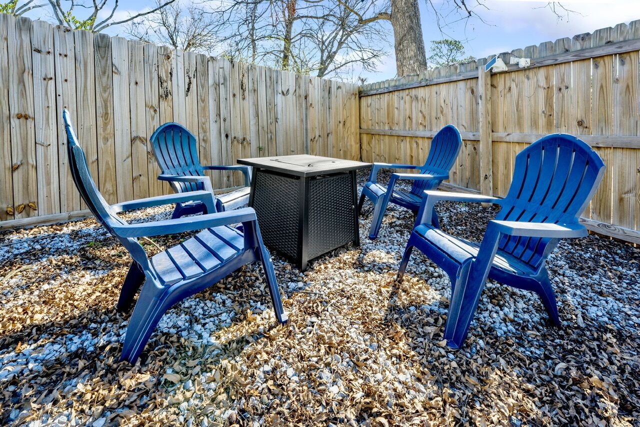 A patio area with a table and four blue chairs, surrounded by a wooden fence and leaf-covered ground.