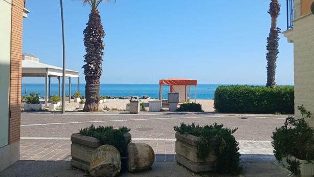 A coastal view with a covered seating area, palm trees, and a building with a balcony. - P085 – Porto Recanati, new two-room apartment 30 meters from the sea (Porto Recanati)