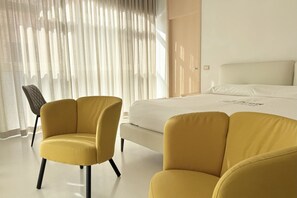 Une chambre d’hôtel avec un lit, deux fauteuils jaunes, une fenêtre avec des rideaux légers et une porte.