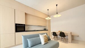 A modern kitchen with a built-in oven, a dining area with a white table and two chairs, and a light blue sofa with a decorative pillow. - P085 – Porto Recanati, new two-room apartment 30 meters from the sea (Porto Recanati)