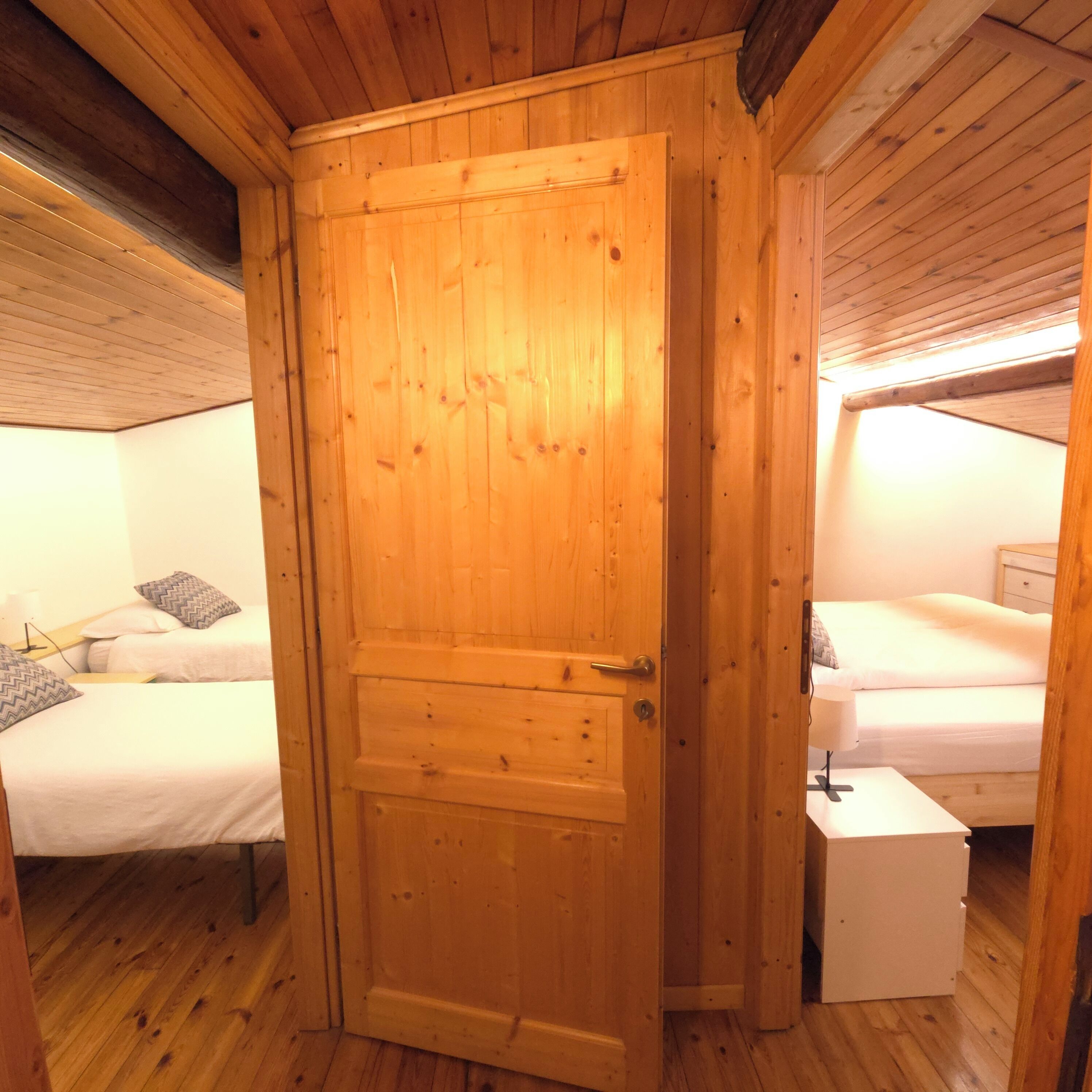A wooden-paneled room with two beds, a white bedside table, and a wooden door.