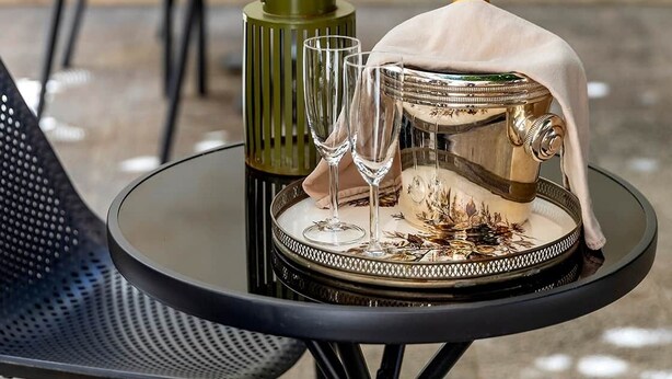 Une table ronde avec un plateau en verre, une bouteille de champagne, deux verres à vin et un plateau décoratif.