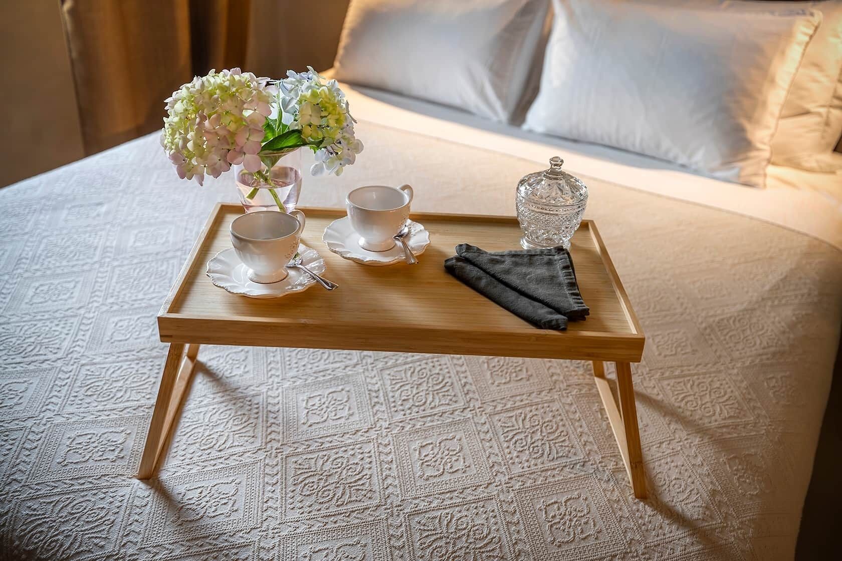 Una bandeja de madera con dos tazas blancas, un vaso y un florero sobre una cama con ropa de cama estampada.