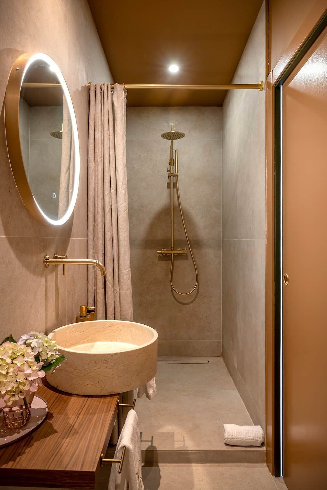 Un baño moderno con un espejo redondo, un mueble de tocador de madera, un lavabo color beige, una ducha con cabezal de ducha manual y una puerta.