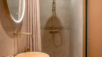 Un baño moderno con un espejo redondo, un mueble de tocador de madera, un lavabo color beige, una ducha con cabezal de ducha manual y una puerta.