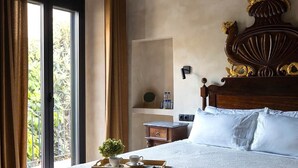 Ein Schlafzimmer mit einem großen Bett, einem Holz-Kopfteil, einem Nachttisch, einer Ablage mit Tasse und Pflanze sowie Blick durch ein Fenster.