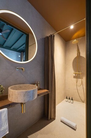 Une salle de bain moderne avec un miroir rond, un lavabo en pierre, une douche équipée d’un pommeau de douche à main et une serviette posée sur le sol.