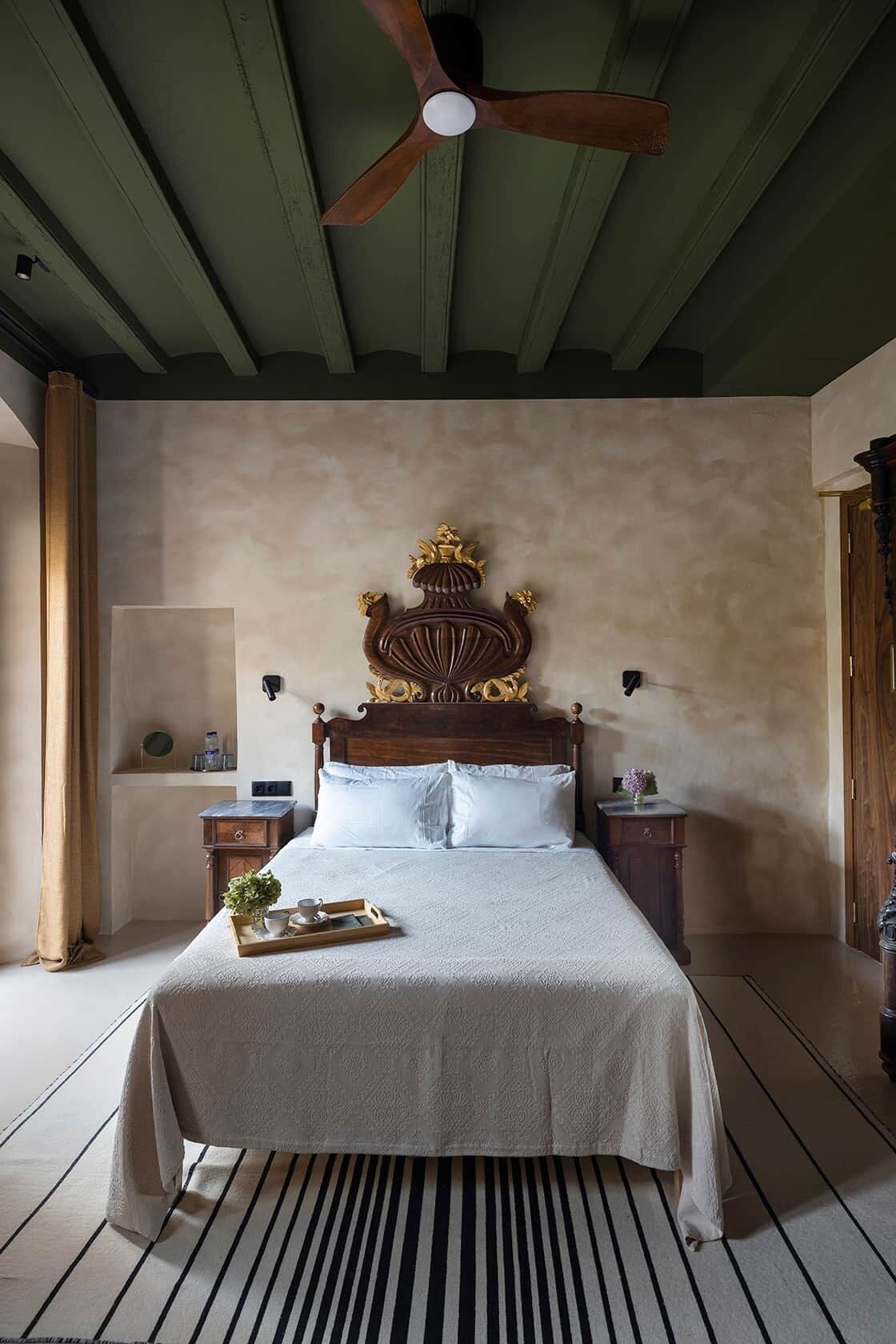 Un dormitorio con cama de madera, ventilador de techo, mesita de noche y bandeja con tazas y platillos.