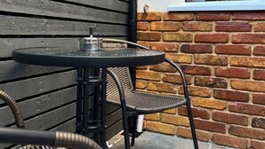 Un patio con una mesa redonda y dos sillas de mimbre, un muro de ladrillo y una ventana que da al interior.
