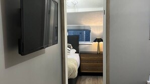 Habitación de hotel con televisor de pantalla plana en la pared, piso de madera, cama con edredón blanco, mesita de noche con lámpara y vistas a un baño a través de un vidrio.