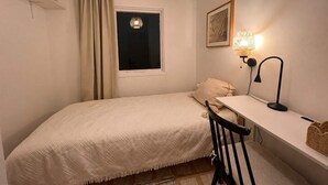 Un dormitorio pequeño y ordenado con una cama individual, un escritorio, una silla, una ventana con cortinas y una lámpara de pared.