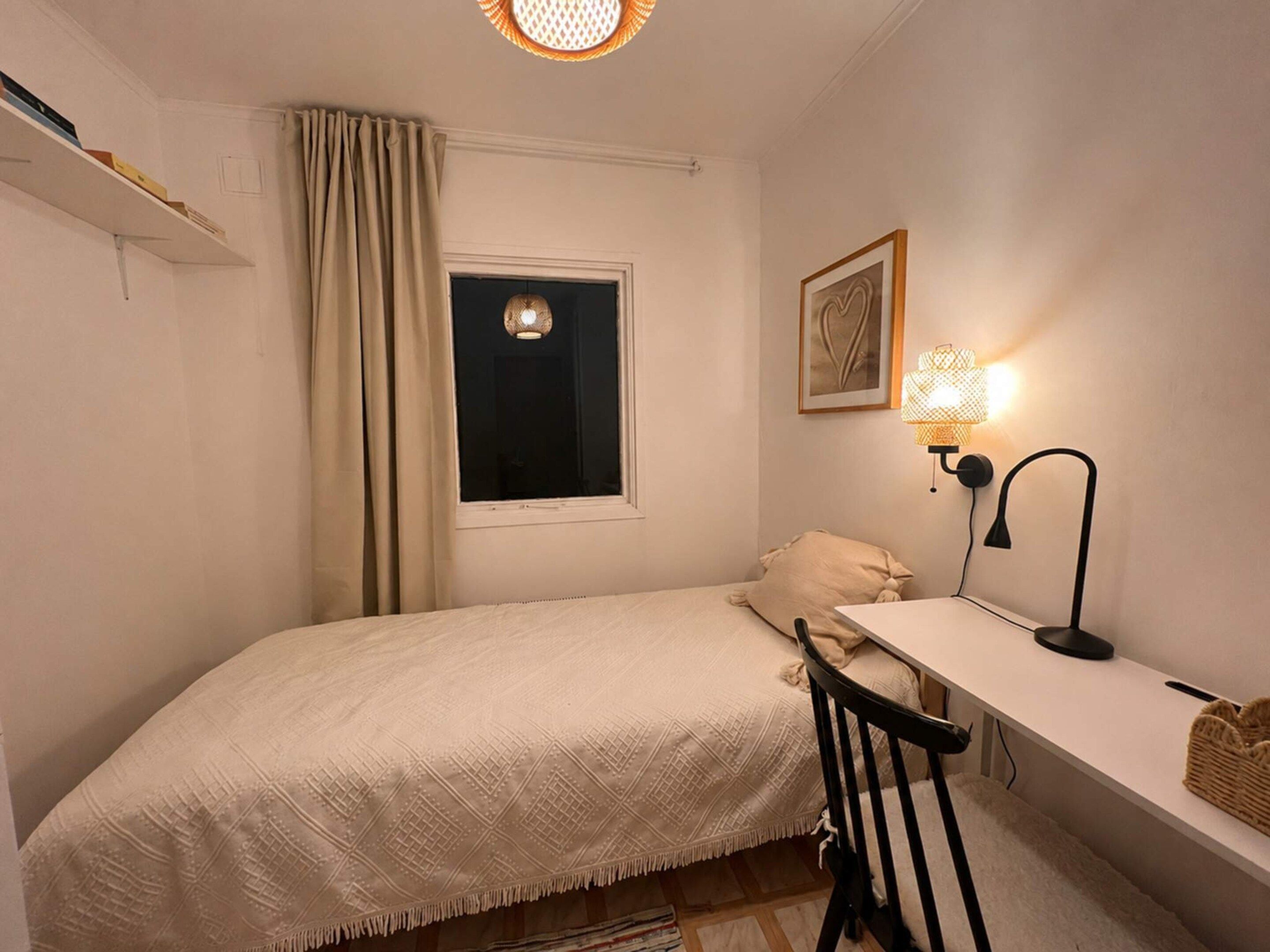 Un dormitorio pequeño y ordenado con una cama individual, un escritorio, una silla, una ventana con cortinas, una lámpara de pared y un cuadro enmarcado.
