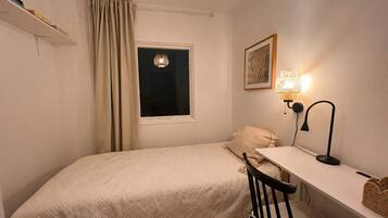 Un dormitorio pequeño y ordenado con una cama individual, un escritorio, una silla, una ventana con cortinas, una lámpara de pared y un cuadro enmarcado.