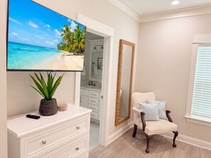 Ein Zimmer mit Fernseher, der eine Strandansicht zeigt, einer weißen Kommode mit Schubladen, einem Sessel mit Kissen, einem großen Spiegel und einer Tür zu einem Badezimmer.