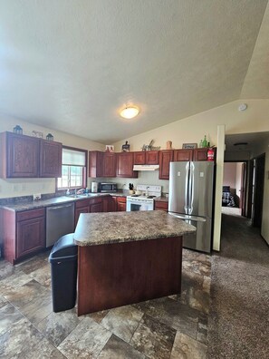 A spacious kitchen with granite countertops, wooden cabinets, a stainless steel refrigerator, and a central island.