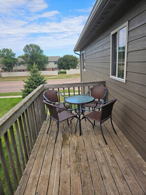 A wooden deck with a round table and chairs, a view of a street, and a house in the background.