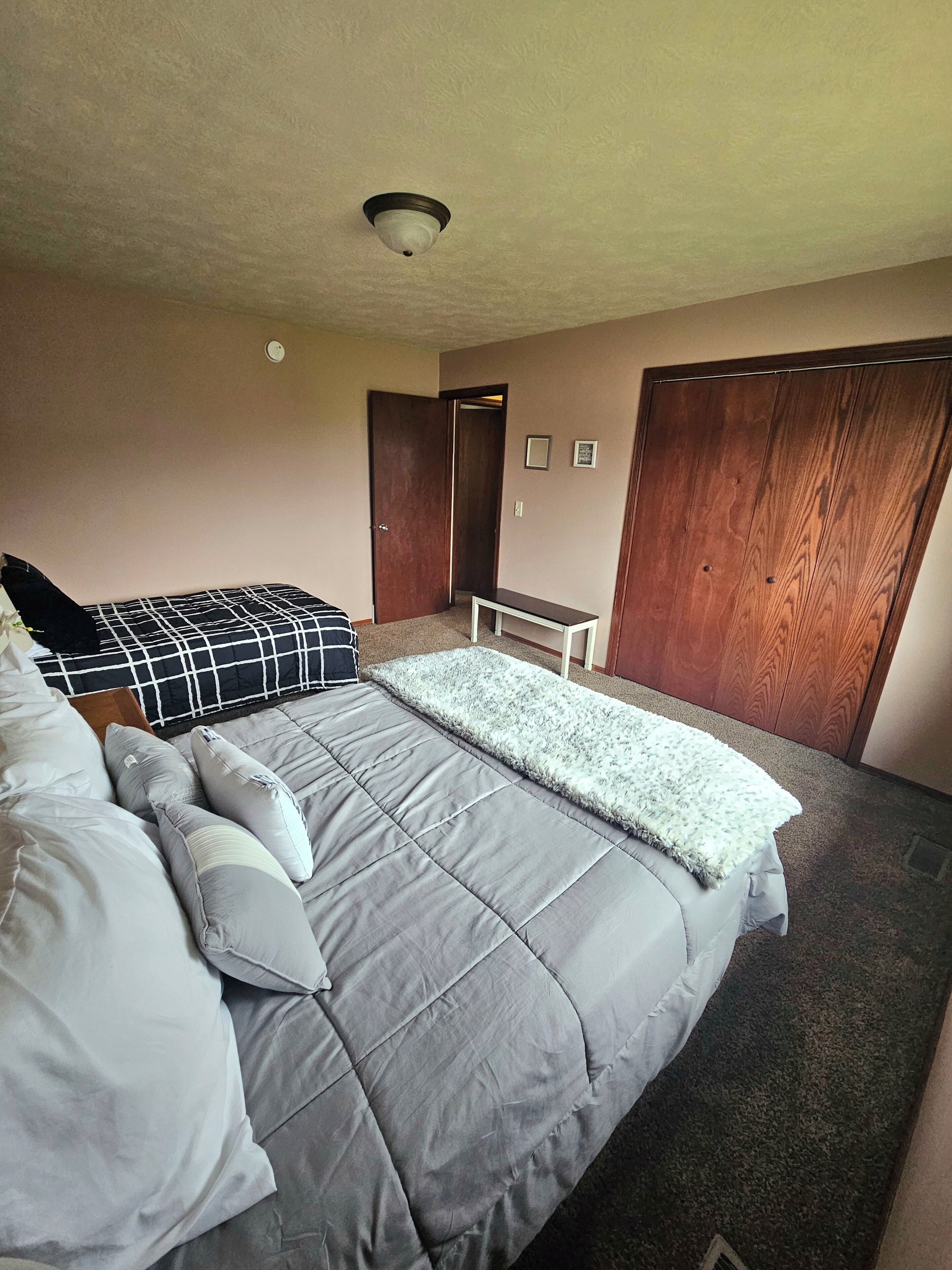 Un dormitorio con una cama, almohadas, una mesita de noche, un armario de madera y una lámpara en el techo.
