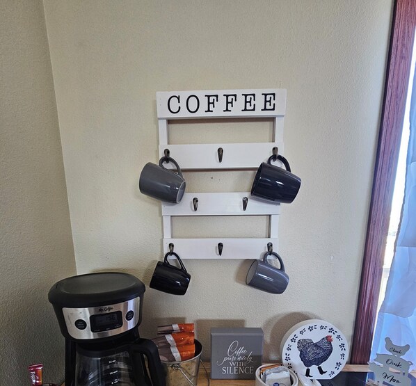 Un coin café avec un panneau, des tasses, une machine à café et une assiette décorée d’un motif de poulet.