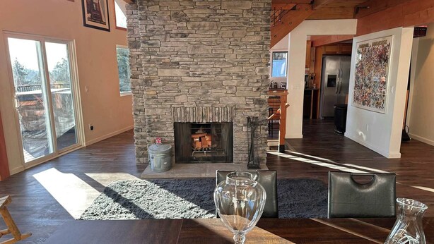 A spacious living room with a stone fireplace, a dining table with a wine glass, and a view of the backyard through large sliding doors. - The Grateful Den - Hot tub, game room (Big Bear Lake)