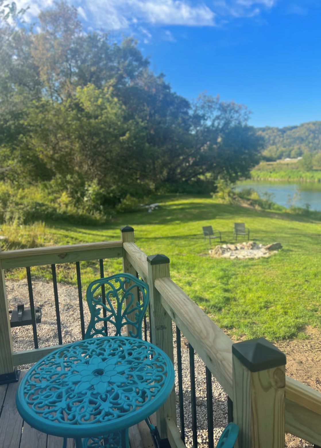A wooden deck with a metal table and chairs, overlooking a grassy area with a pond and trees.
