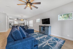 A modern living room with a blue sofa, a ceiling fan, a flat-screen TV, and a large window.