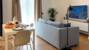 A modern living room with a grey sofa, dining table, and a TV on a white cabinet. - April Suites Apartments - Suite 1, Amber (Palermo)