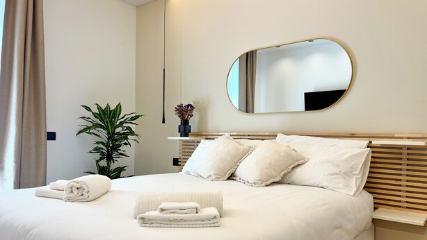 A neatly made bed with pillows, a bedside table with a vase of flowers, a large mirror on the wall, and a potted plant. - April Suites Apartments - Suite 1, Amber (Palermo)