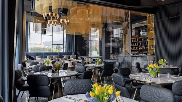 A modern restaurant with round tables, black upholstered chairs, and a bar area visible through a glass partition. - Hotel Cube (Bar)