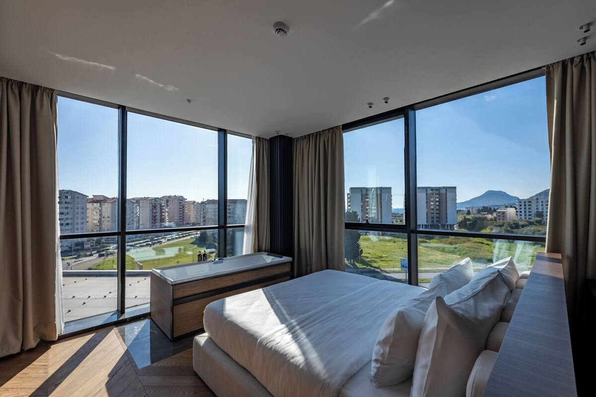 A modern hotel room with large windows, a bed, a bench, and a view of the cityscape.