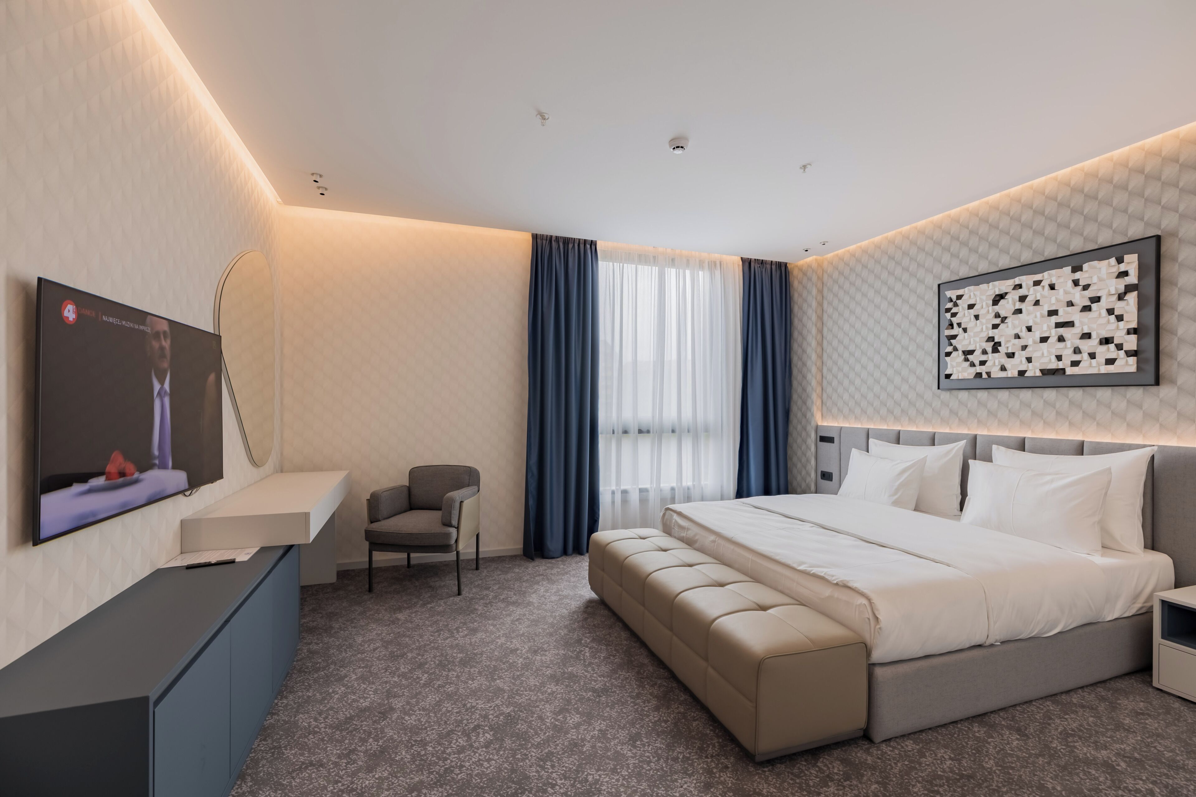Quarto de hotel moderno com cama, escrivaninha, cadeira e televisão.