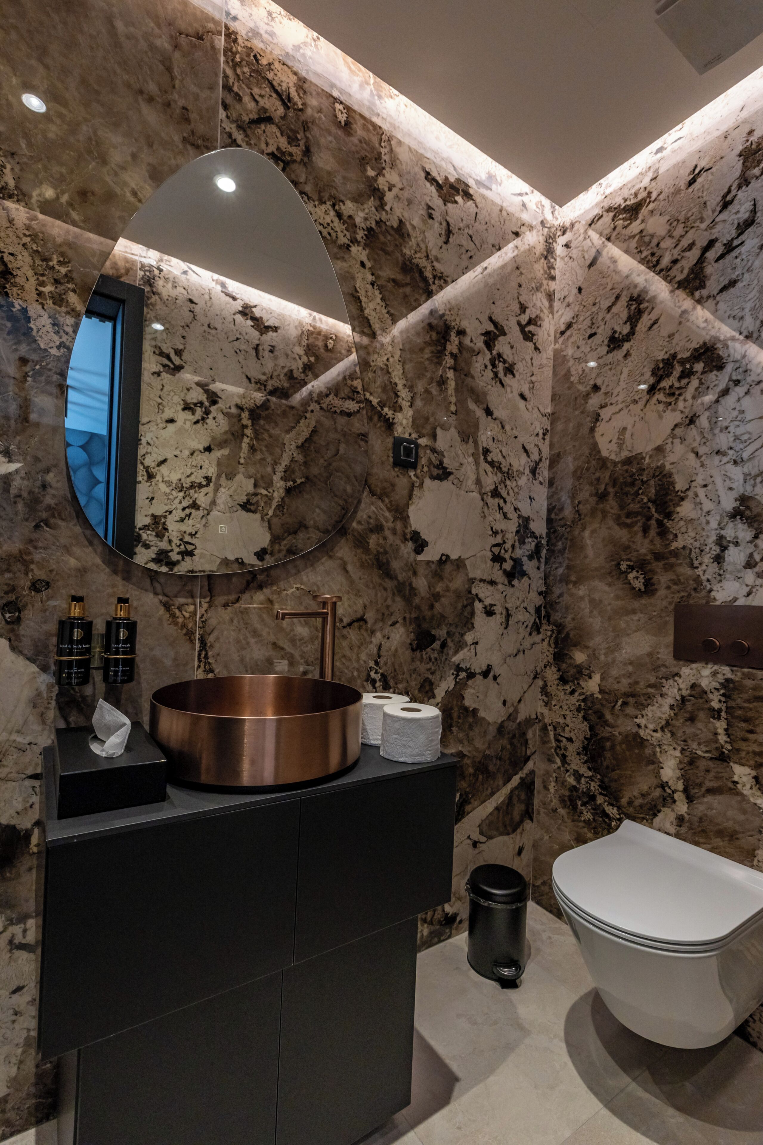 A bathroom with a marble wall, a round mirror, a copper sink, a toilet, and a trash bin.