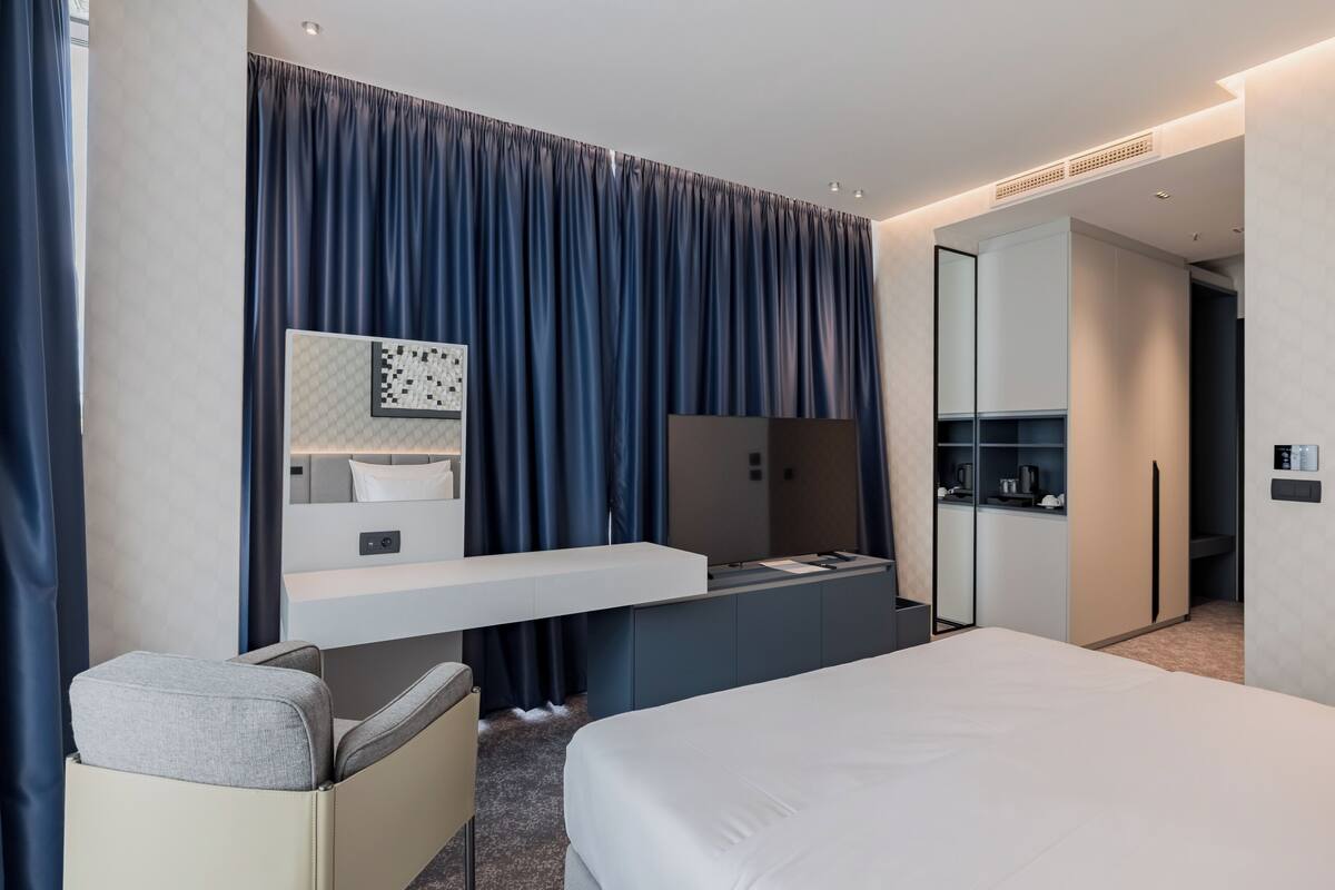 A modern hotel room with a bed, a desk, a chair, a large mirror, and a wardrobe.