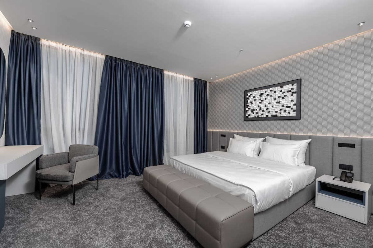 A modern hotel room with a bed, bedside table, chair, and a large window with curtains.